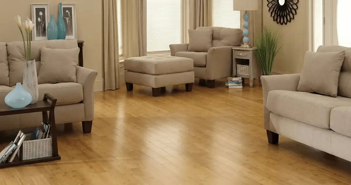 A living room with professionally installed bamboo flooring, beige sofas, a matching ottoman, a side table with a blue lamp, vases, framed pictures, and large windows with blinds and curtains in Saint Louis, MO